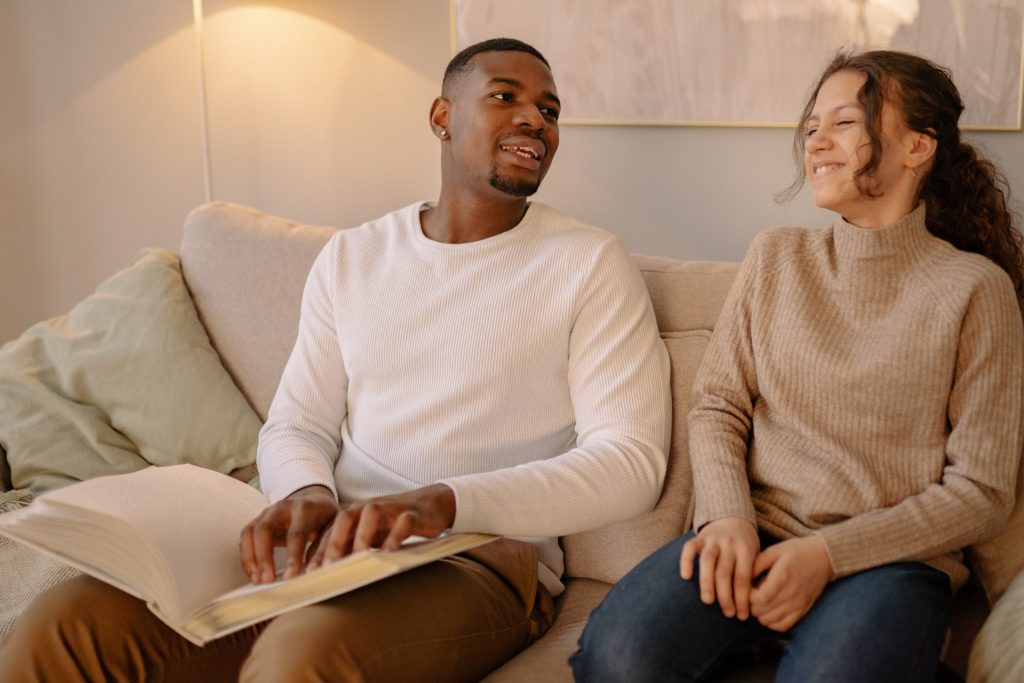 Visually impaired man reading a braille book on a couch, smiling and engaging with a woman sitting next to him