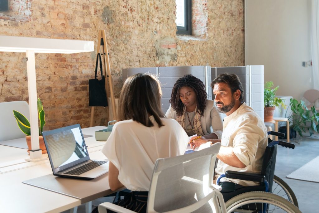 Diverse team meeting in a modern, accessible office: a man in a wheelchair, a woman, and a third colleague collaborating at a large desk with a laptop.