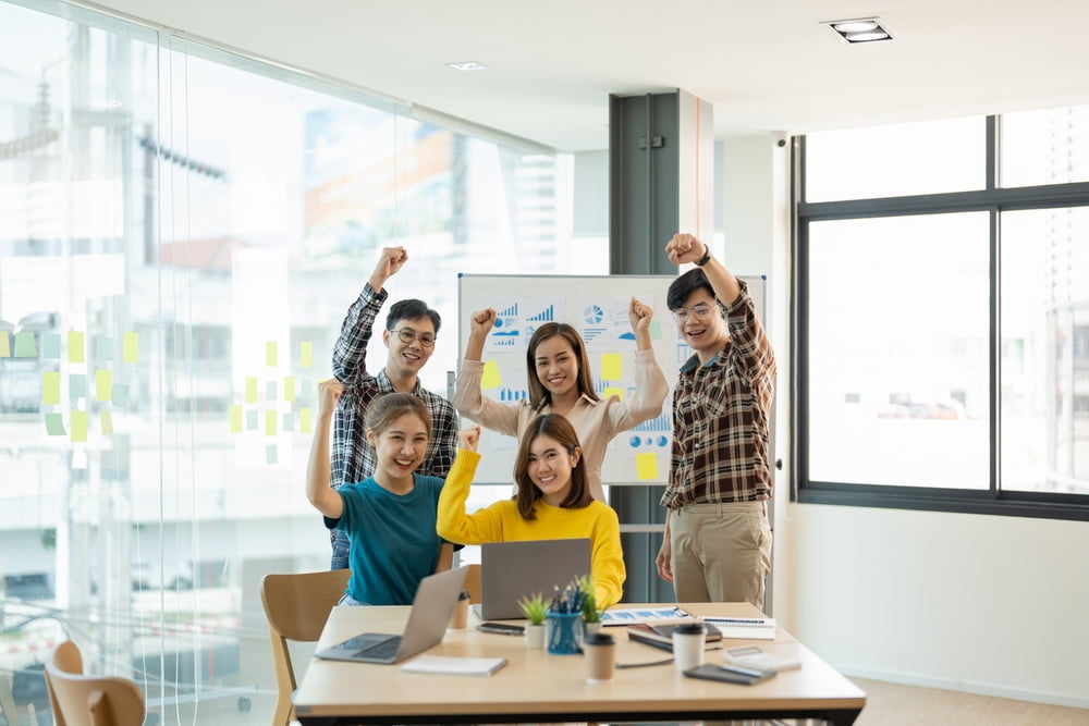 Diverse, happy team of five employees celebrating success with raised fists in a bright, modern, accessible office.