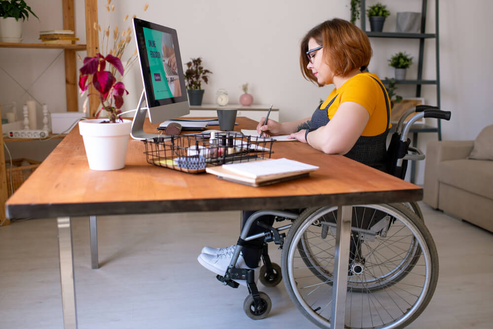 A woman in a wheelchair is seated at a desk, focused on her work with a laptop in front of her.