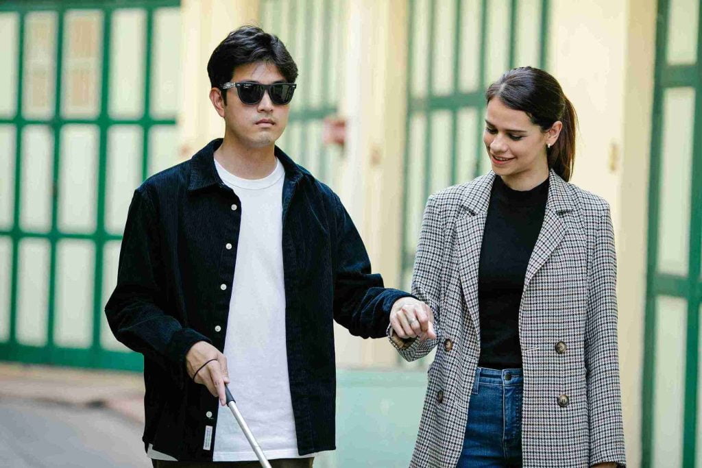 Visually impaired man with a white cane, guided by a female companion, walking outdoors.