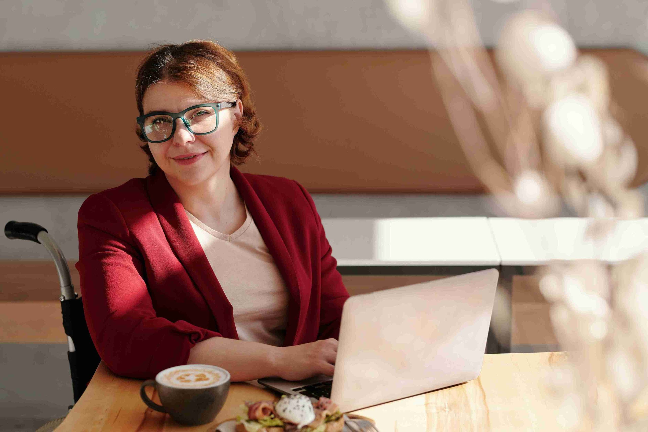 A woman in a wheelchair wearing a red jacket and glasses, smiling while working on a laptop at a cafe table.