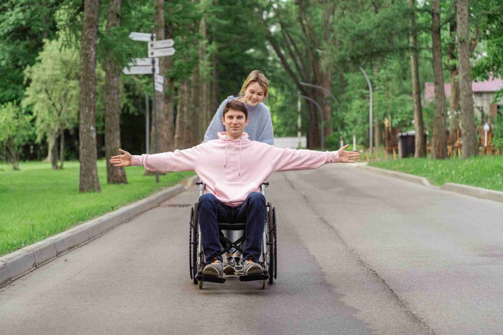 Happy young man in a wheelchair with arms outstretched, being pushed by a friend or caregiver along a tree-lined path.