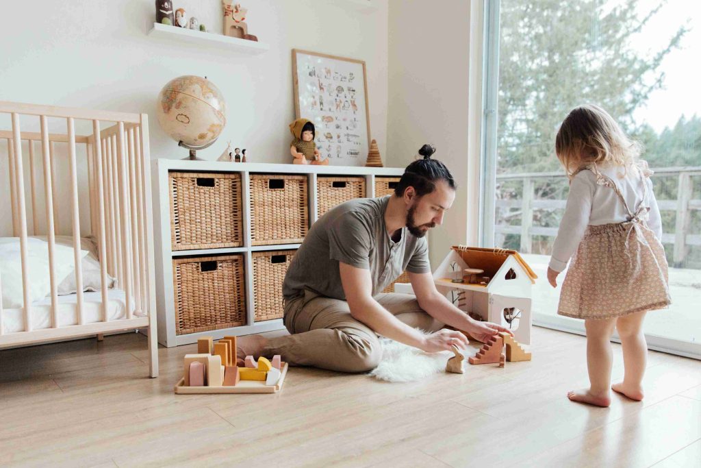 "Caring father sitting on the floor of a bright nursery, playing with wooden blocks and his young toddler daughter