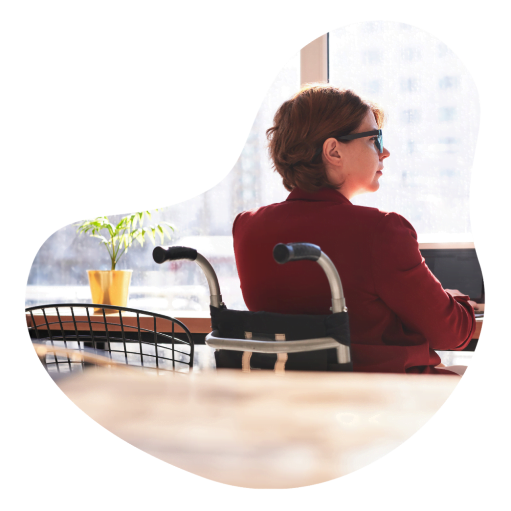 A woman in a wheelchair is seated at a table, working on her laptop with a focused expression.