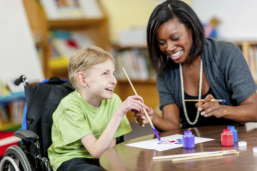 Happy boy in a wheelchair smiling while painting with a female support worker or art therapist.